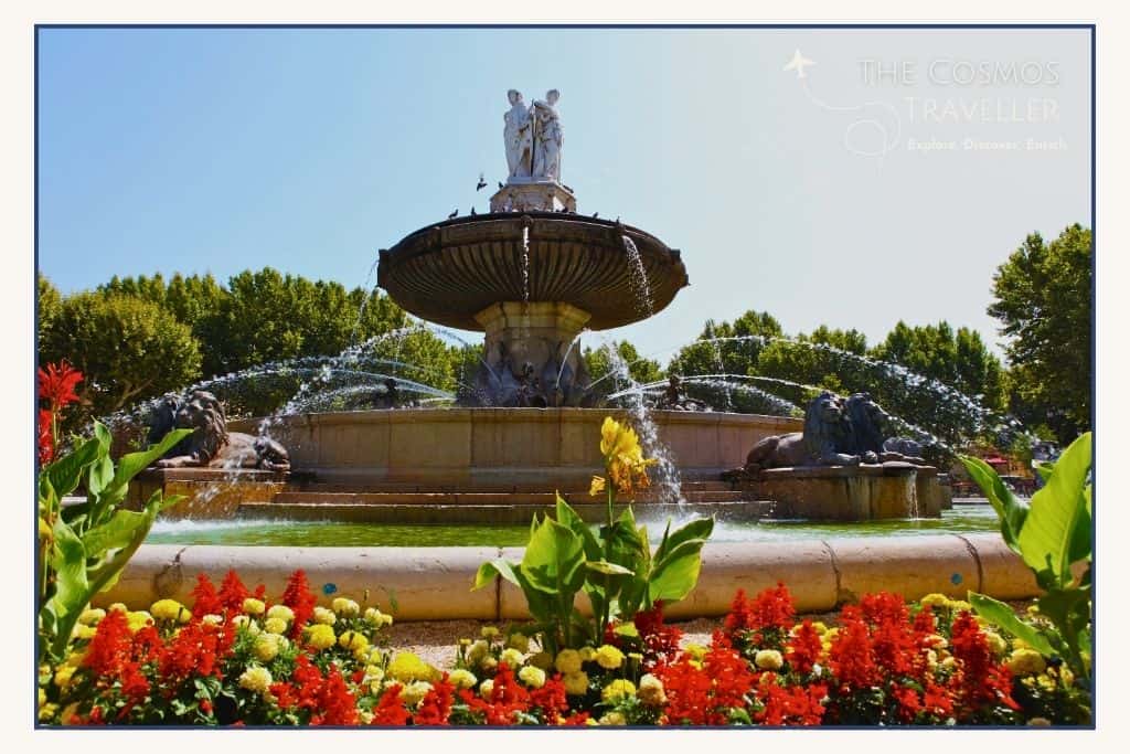 The Fontaine de la Rotonde in Aix-en-Provence, surrounded by vibrant flowers, lion sculptures, and plane trees on a sunny day.