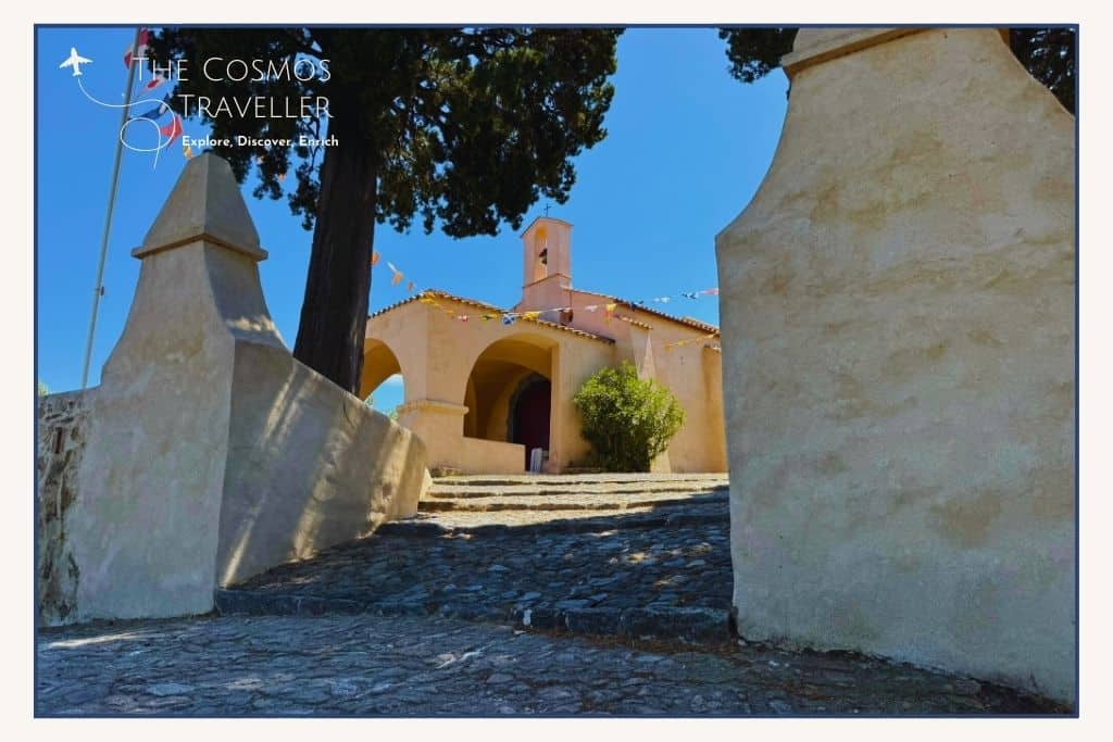 Pastel-coloured Chapel of Sainte-Anne in Saint-Tropez with flags fluttering under a bright blue sky, seen from the entrance.