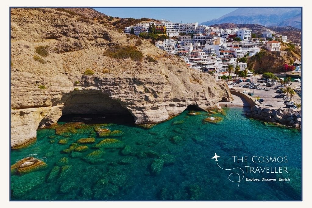 Drone shot of Agia Galini beach with turquoise water, cliffs, and resort buildings in the background.