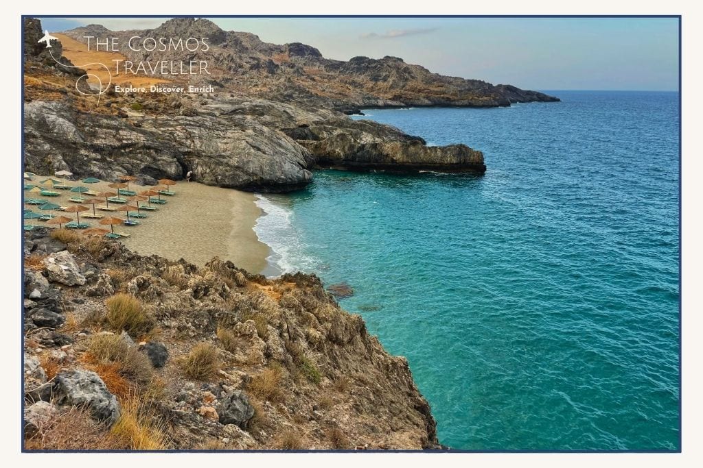 Mikro Ammoudi Beach in Crete at morning light, with sun loungers on sandy shore and rocky coast in background.