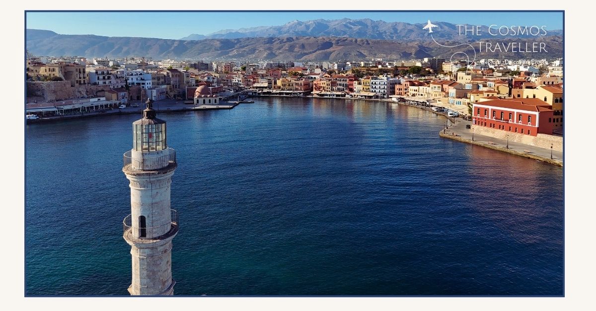 Drone view of Chania lighthouse and colorful harbour with white mountains in the background.