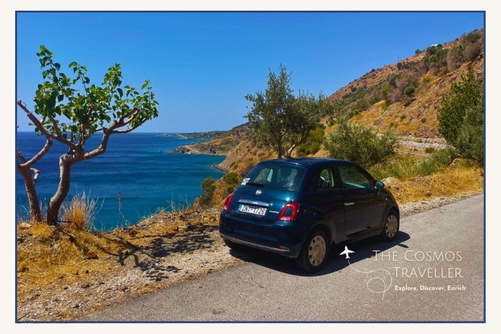 A blue Fiat 500 hire car parked on a winding coastal road in Crete with sea views in the background.