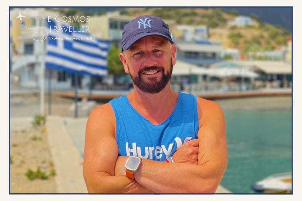 The Cosmos Traveller founder in Greece, smiling with cap and beard, Greek flag behind — Crete travel expert.