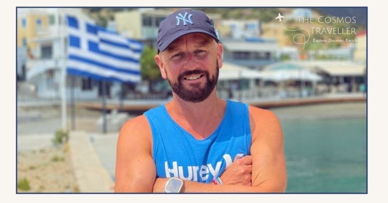 The Cosmos Traveller founder in Greece, smiling with cap and beard, Greek flag behind — Crete travel expert.